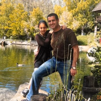 A man and a woman posing together on a rock by a pond in a park, with a wooden observation tower in the background.