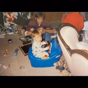 Child in toy car with man A young child in pajamas sits in a blue Little Tikes ride-on toy car on a carpeted floor, while a man in a plaid shirt sits behind him. The room is filled with Christmas decorations and scattered toys.