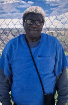 Portrait of Verne, an older man with glasses and a blue shirt, smiling in front of a chain-link fence overlooking a valley.