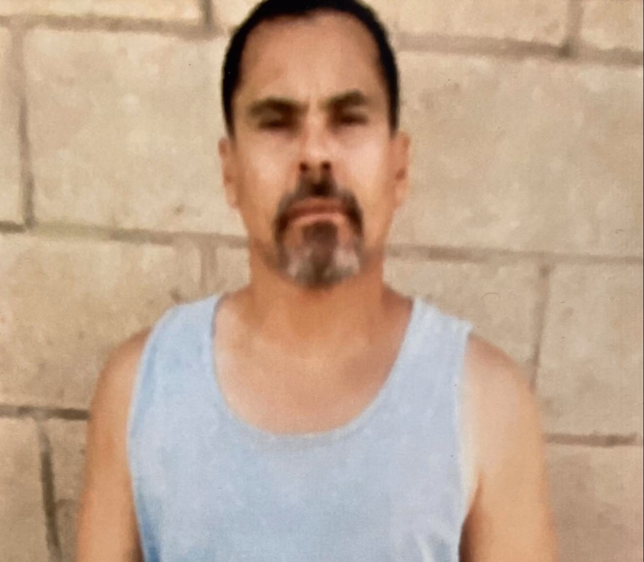Man with a goatee wearing a light blue tank top against a light-colored brick wall.