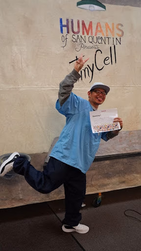 A man in a blue shirt and cap poses energetically in front of a wall featuring the 'HUMANS of SAN QUENTIN Presents JimmyCell' logo.