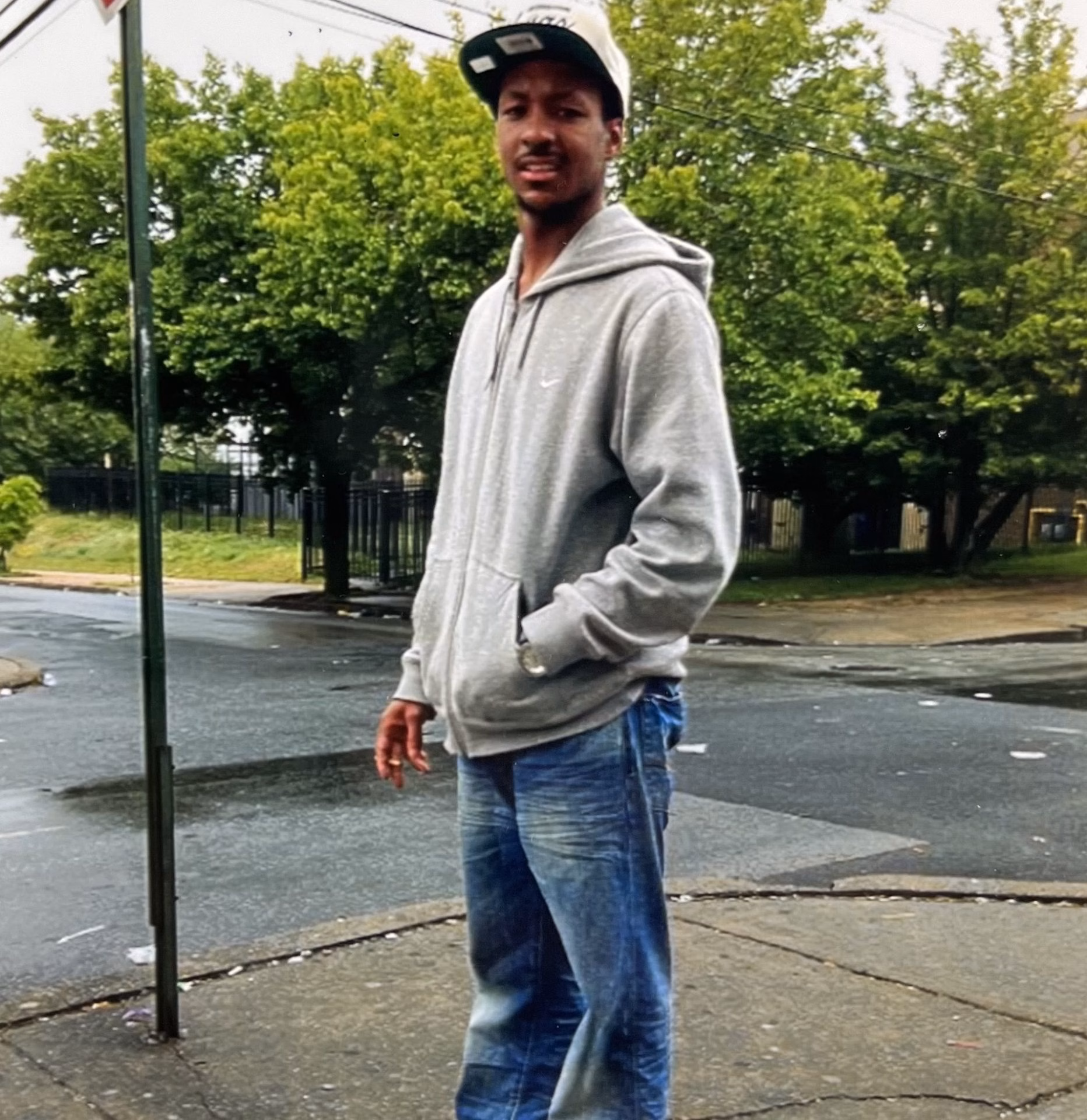 A young man wearing a grey Nike hoodie and blue jeans stands on a sidewalk, with a green pole and trees in the background.