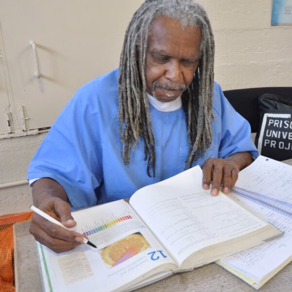 Eddie Herena, a man with long grey dreadlocks in a blue shirt, studies an open textbook while holding a white pen.