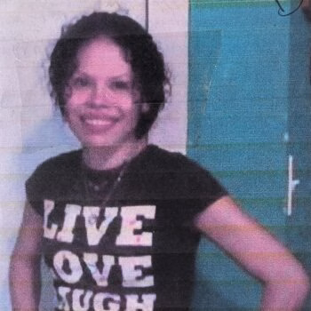 A smiling woman with dark curly hair wearing a black t-shirt that says 'LIVE LOVE HUGH' in white block letters.