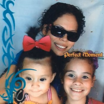 A woman wearing sunglasses smiles above two young girls, one with a red bow and the other with a purple flower, against a blue background with decorative swirls and the text 'Perfect Moment'.