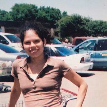 A woman with dark hair smiling in a parking lot with cars and trees in the background.