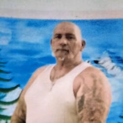A bald man with a goatee and tattoos on his arms, wearing a white tank top, stands in front of a blue background.