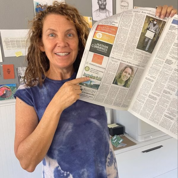 A woman in a blue tie-dye shirt holds up a newspaper, pointing to an article about the Nathan Cummings Foundation and Diann Kahn.