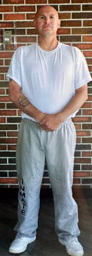 A bald man in a white t-shirt and grey sweatpants stands against a brick wall.