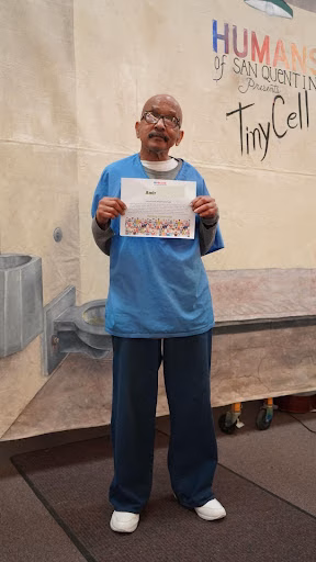 Amir, 73, wearing a blue scrub top and holding a certificate in front of a mural for the Humans of San Quentin Foundation TinyCell.