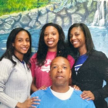 A man in a blue shirt sits in front of three smiling women, all posing together in front of a painted waterfall backdrop.