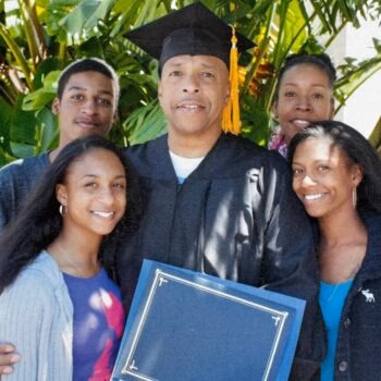 A family of five poses for a graduation photo. A man in a black cap and gown holds a blue diploma frame, surrounded by four smiling family members outdoors.