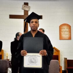 A man in a black graduation cap and gown holds a framed Certificate of Achievement for Todd Williams in front of a church cross.