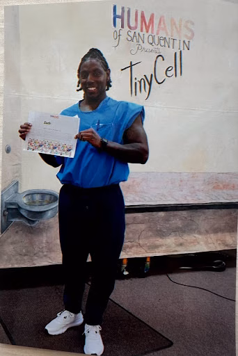Zach R. stands in front of a promotional backdrop for the Humans of San Quentin event, holding a certificate.