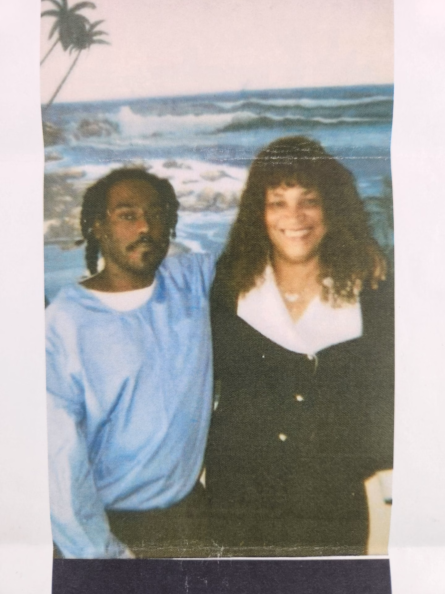A man with dreadlocks and a woman with curly hair smiling together in front of a beach backdrop.