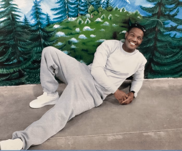 A smiling man in a white long-sleeved shirt and grey sweatpants lies on a concrete floor in front of a painted forest mural.