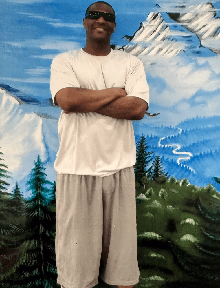A man wearing sunglasses and a white t-shirt stands with arms crossed in front of a painted mountain landscape.