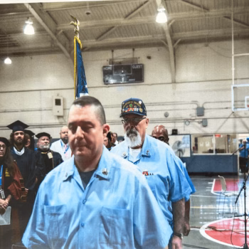 Two men in light blue shirts walk through a gymnasium during a ceremony, with graduates in caps and a flag in the background.