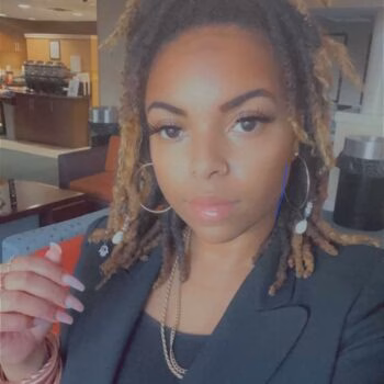A woman with braided hair and large hoop earrings wearing a black blazer, looking at the camera.