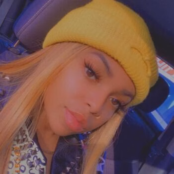 A close-up selfie of a woman with long blonde hair wearing a yellow beanie and a nose ring, sitting inside a car.