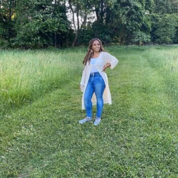 A woman with long braids stands in a green field, wearing a white cardigan, blue jeans, and sneakers, with her hand on her hip.