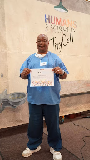 Carlos, 57, holds a certificate in front of a 'Humans of San Quentin' backdrop.