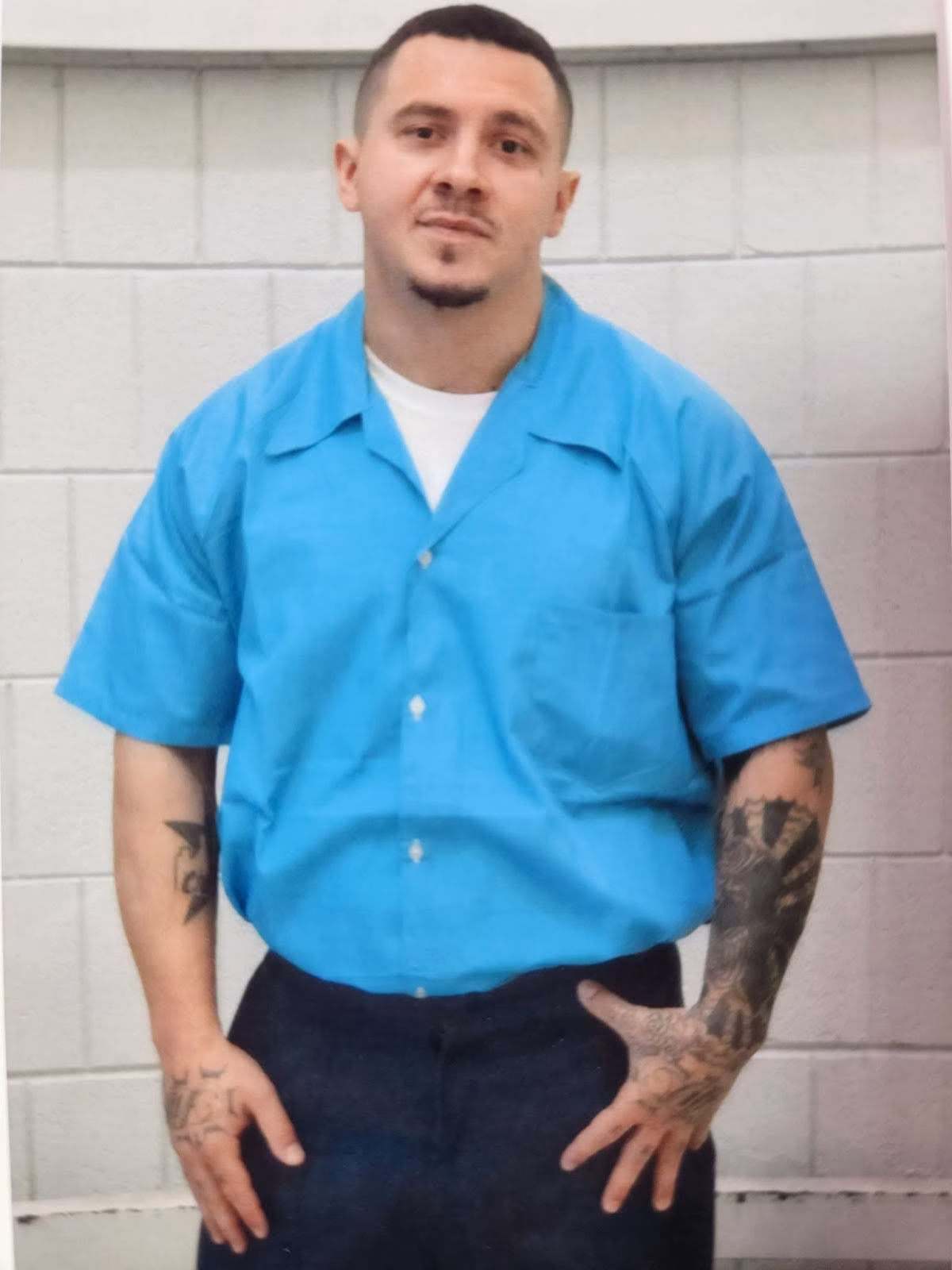A man with tattoos on both arms stands in a blue prison uniform against a white cinder block wall.