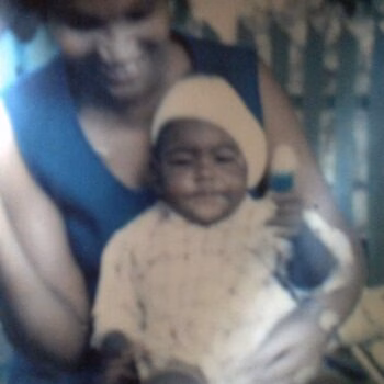 Woman holding baby A woman in a blue top holds a smiling baby wearing a light-colored hat and patterned shirt.
