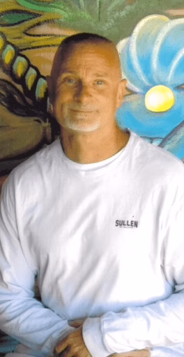A man in a white Sullen long-sleeved shirt stands in front of a colorful mural.