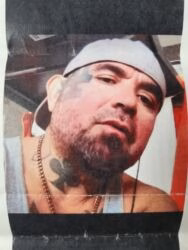 A man with a beard and a white cap, wearing a necklace and a chest tattoo of a club.