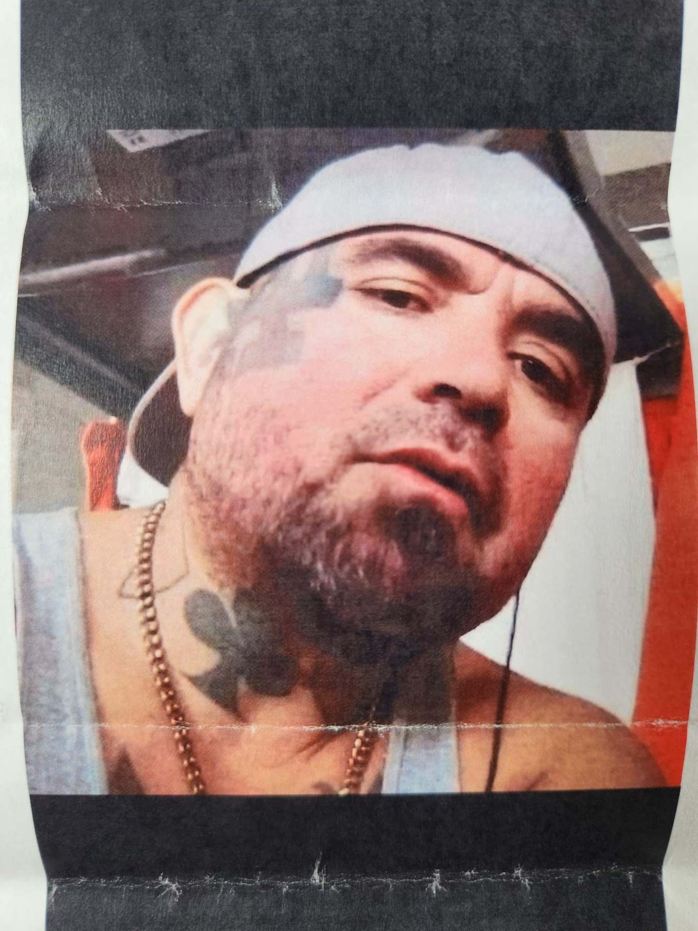 A man with a beard and a white cap, wearing a necklace and a chest tattoo of a club.