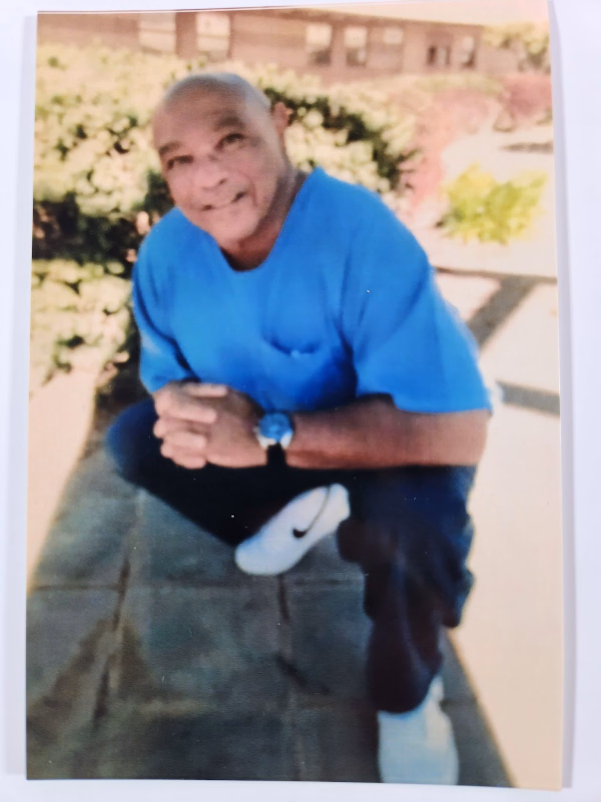 Anthony Trotter, a bald man in a blue shirt and dark pants, smiles while crouching on a stone patio with a building and greenery in the background.