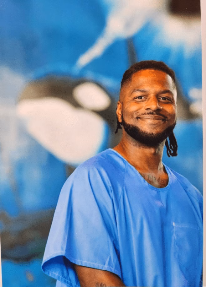 A man in blue scrubs smiles in front of an orca mural.