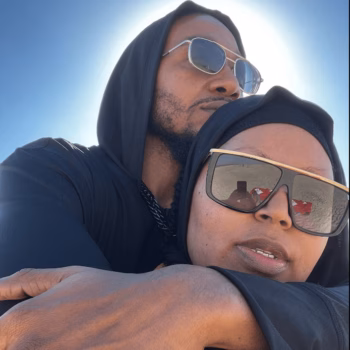 Couple embracing outdoors A man and a woman in black hoodies and sunglasses embrace outdoors under a bright sun.