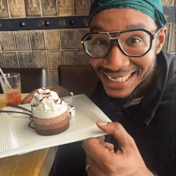 Man with dessert A man wearing a green headwrap and glasses smiles while holding a white plate with a layered chocolate and whipped cream dessert.