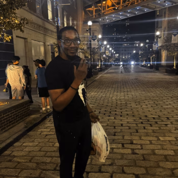 Man on Cobblestone Street A man in a black t-shirt and glasses stands on a cobblestone street at night, holding a white bag and gesturing with his hand.