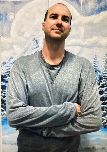 Cameron, a bald man with a beard, stands with his arms crossed in front of a winter landscape mural.