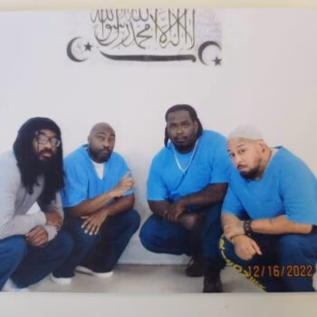 Four men in blue shirts crouching against a wall with Islamic calligraphy above them.