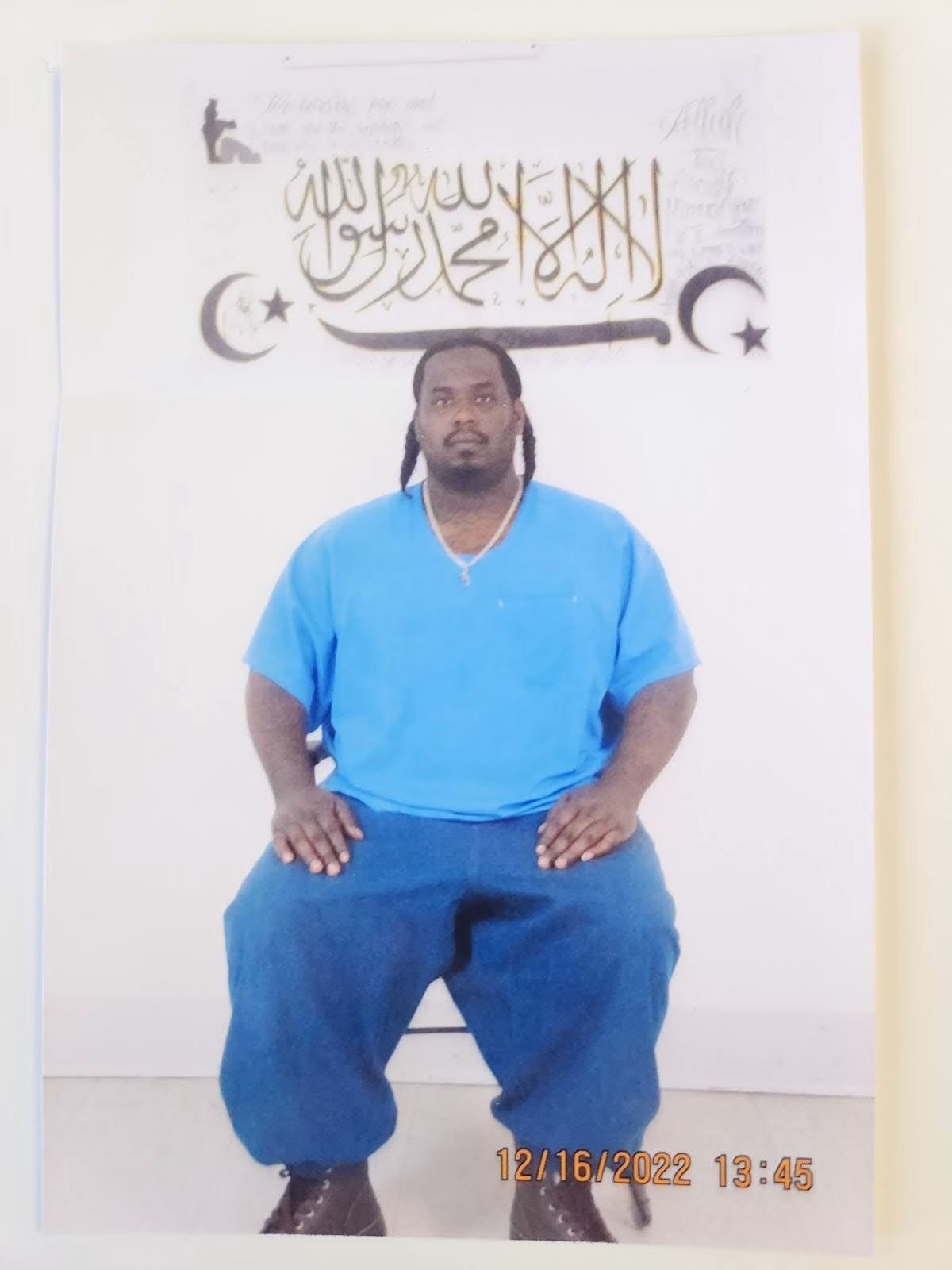 A man with braids in a blue shirt and baggy pants sits in front of a wall decorated with Islamic calligraphy and crescent stars.