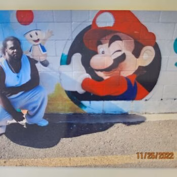 A man in a white tank top and pants poses in front of a colorful mural featuring Mario, Toad, and Yoshi on a brick wall.