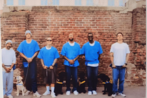 Six men, five in blue prison uniforms and one in a white t-shirt, stand in a row with their service dogs against a brick wall.