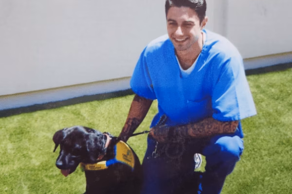 A smiling man in blue scrubs kneels on grass next to a black service dog wearing a yellow vest.