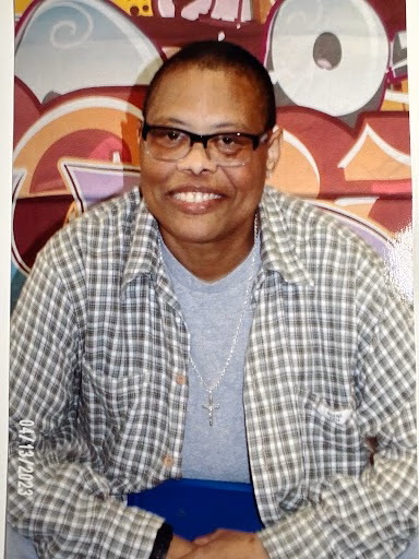 Carol, a smiling woman with glasses, wearing a plaid shirt over a grey t-shirt and a cross necklace, sits in front of a colorful, abstract background.
