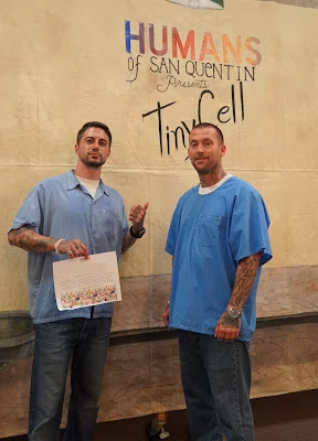 Two men in blue scrubs stand before a sign for 'Humans of San Quentin Presents Tim Cell'.