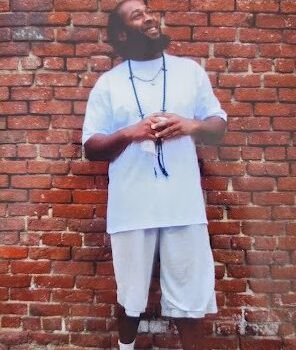 jonquil A man with a beard and glasses stands against a red brick wall, wearing a white t-shirt, shorts, and sneakers.