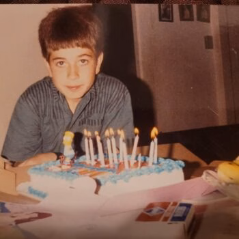 A young boy with brown hair in a striped shirt sits behind a birthday cake with lit candles and a Homer Simpson figurine.