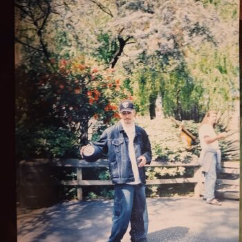 A man named jose3 stands in a park, wearing a denim jacket and cap, with another person in the background.
