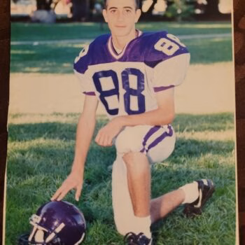 A young man in a purple and white football uniform with the number 88 kneels on a grassy field, his hand resting on a purple helmet.