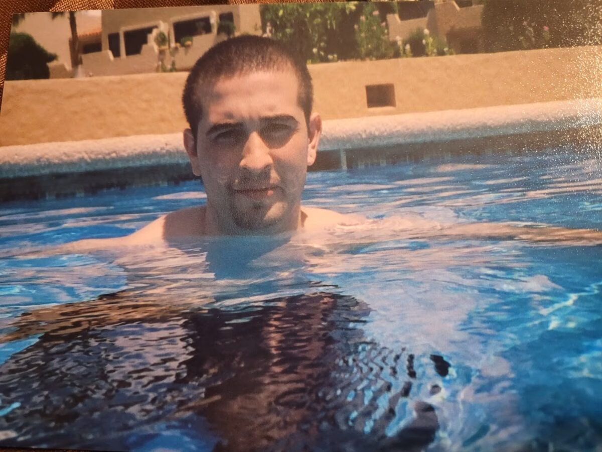 A man with short dark hair and a goatee is submerged in a swimming pool up to his chest, looking directly at the camera.