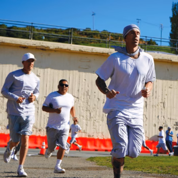 Three men in white athletic wear run on a gravel path under a clear blue sky, with a concrete wall and green hills in the background.